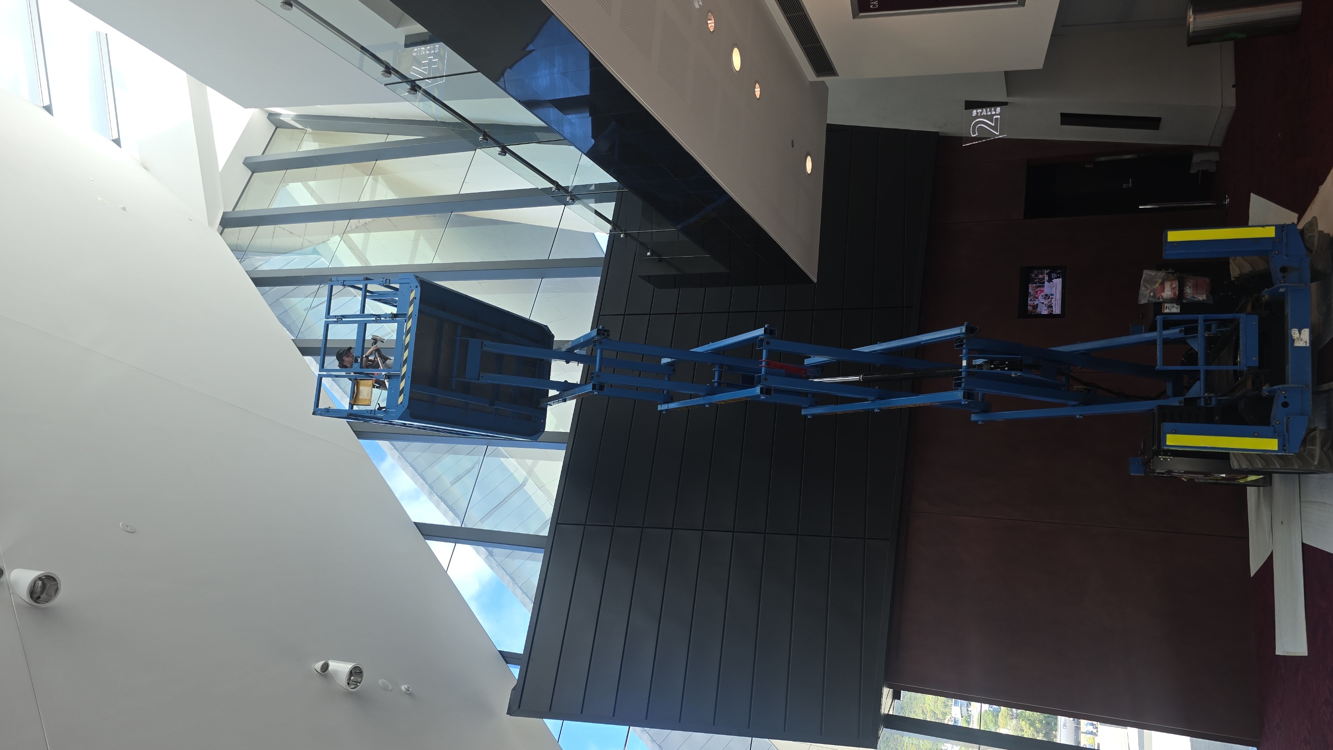 A person is working on a scissor lift inside a modern building with large windows and high ceilings.