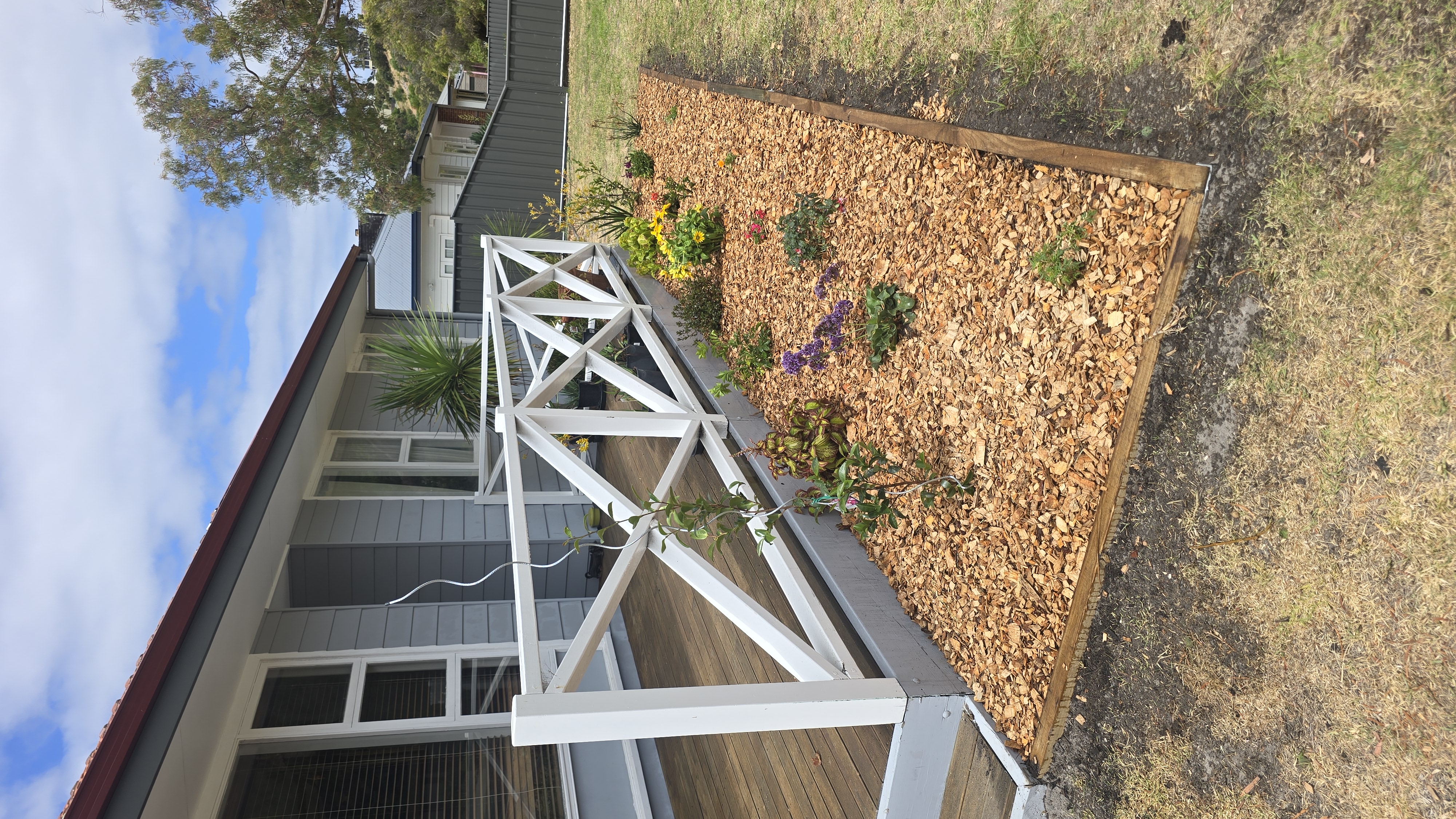 A small front garden with a wooden border, covered with wood chips, and planted with various small flowers. A wooden deck with a white railing extends from a gray house with large windows. A large potted plant with long, pointed leaves is on the deck. A wire is hanging from the railing. The background features a cloudy sky and neighboring houses with fences.