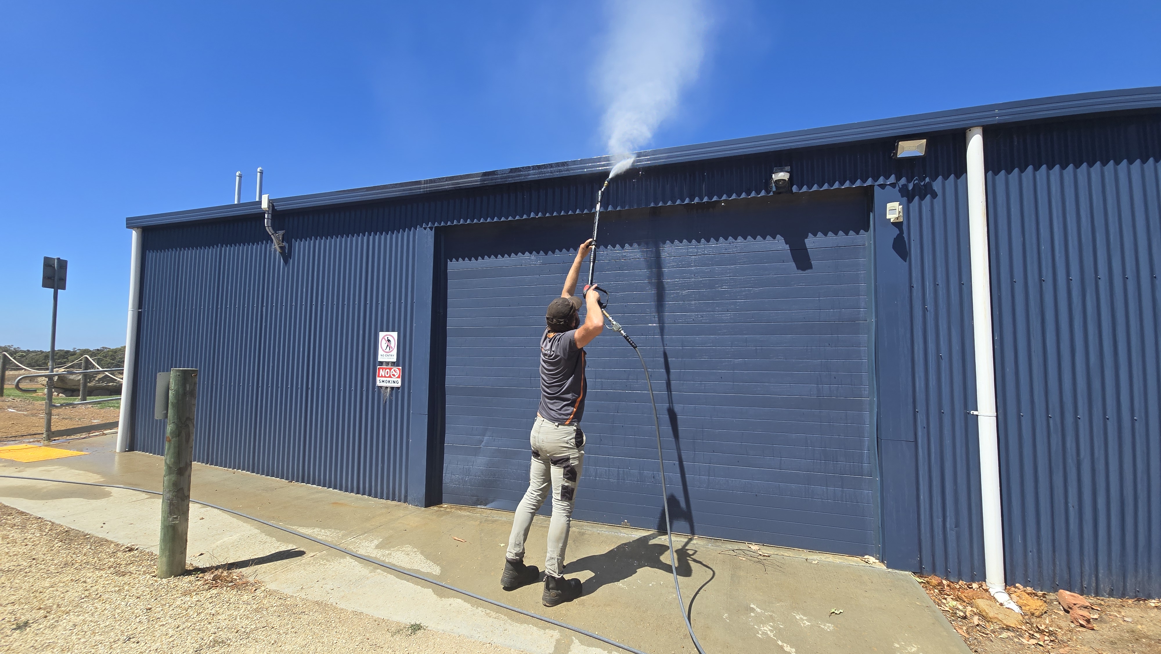 Person power washing a blue building exterior under clear blue sky.