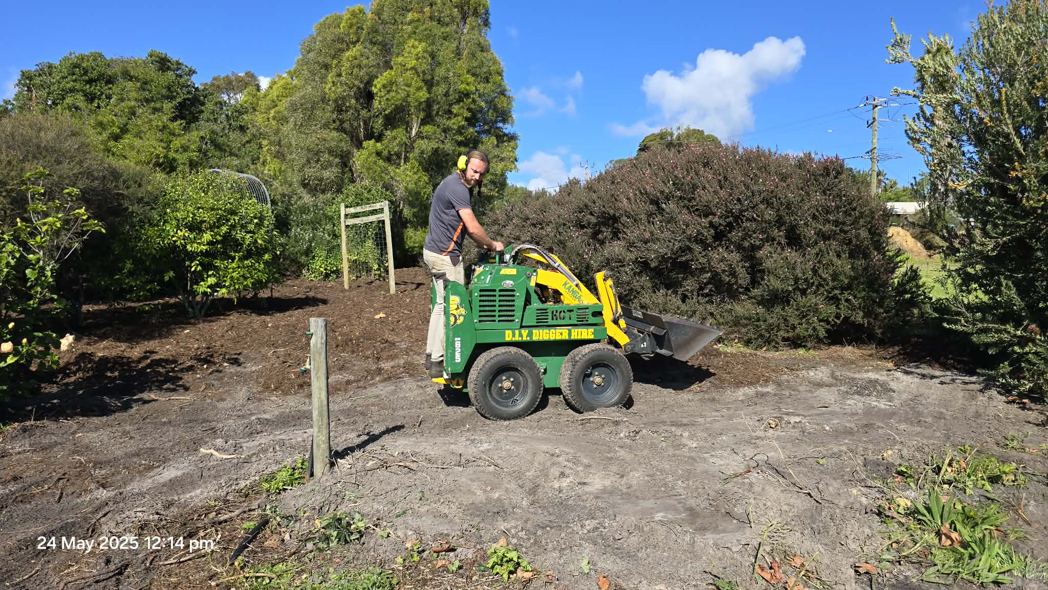 A man operating a small green and yellow industrial ground cleaning machine labeled 'D.I.Y. Digger Hire' in a garden area with trees, shrubs, a wooden fence, and a blue sky with clouds.
