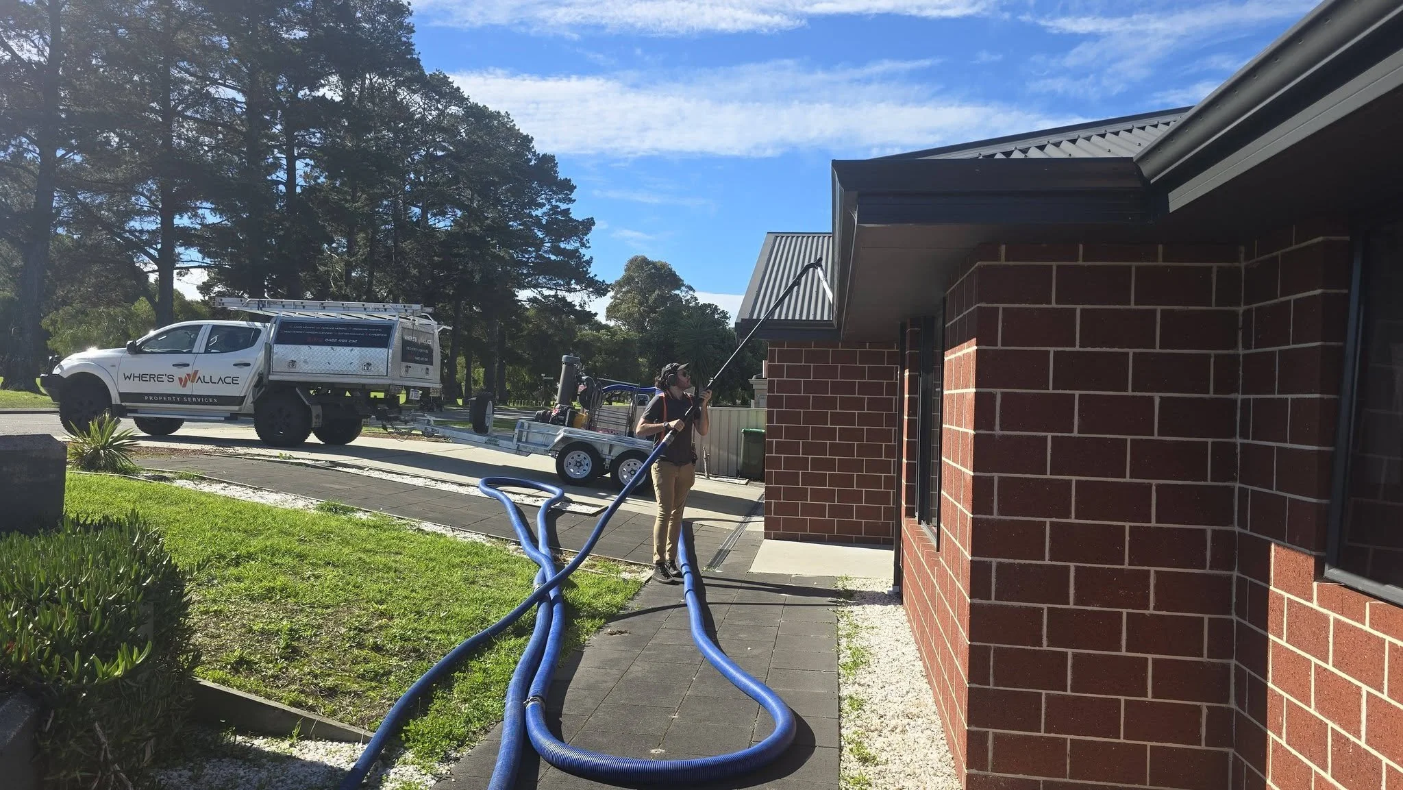 A worker using a long pole to spray or clean the exterior wall of a red brick building, with a white service truck labeled "WHERE'S WALLACE PROPERTY SERVICES" parked on the street nearby and supplies on a trailer, under a blue sky with scattered clouds.