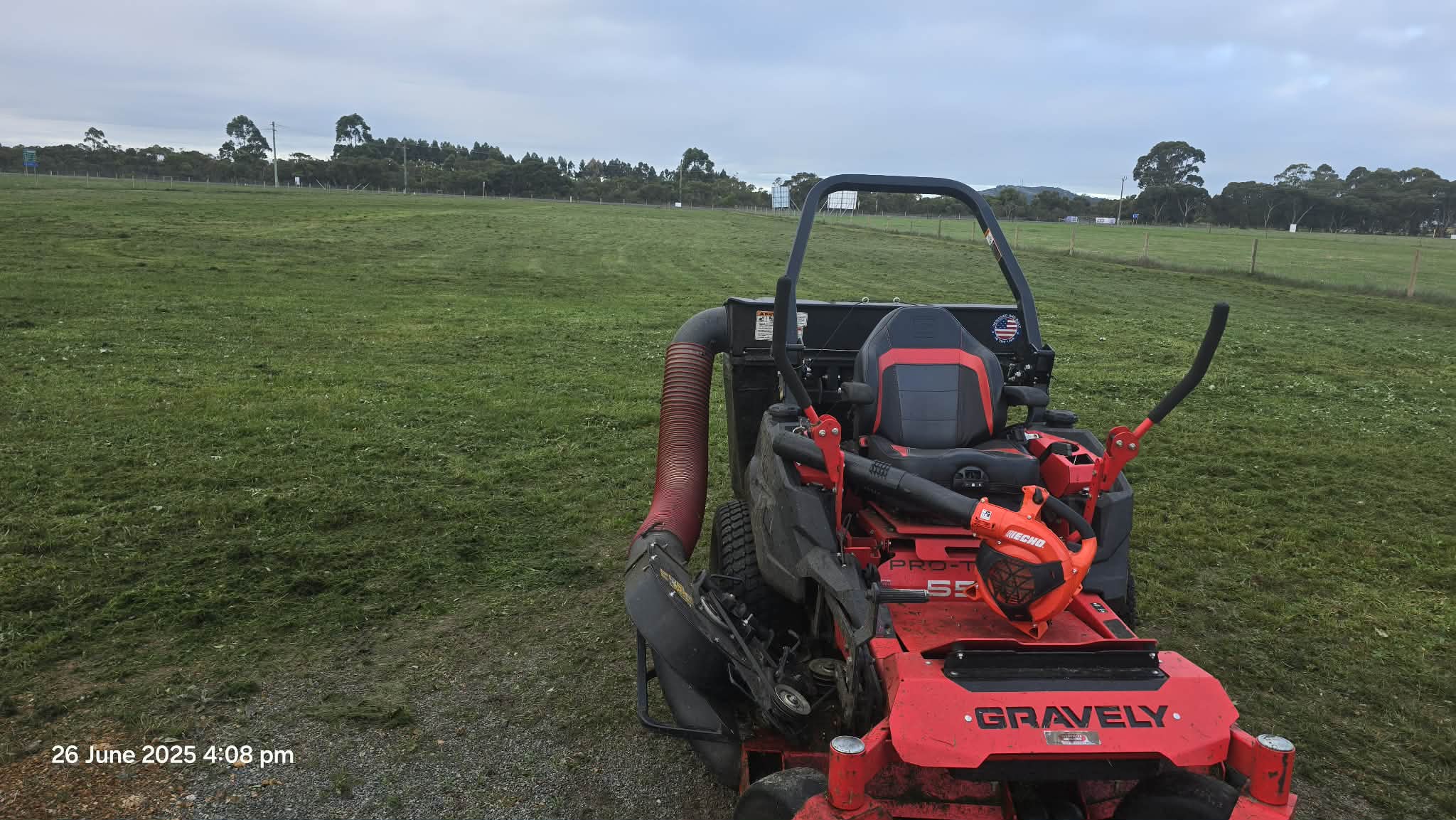 A red and black Gravely lawn mower on a grassy field with a few trees in the background under a cloudy sky.