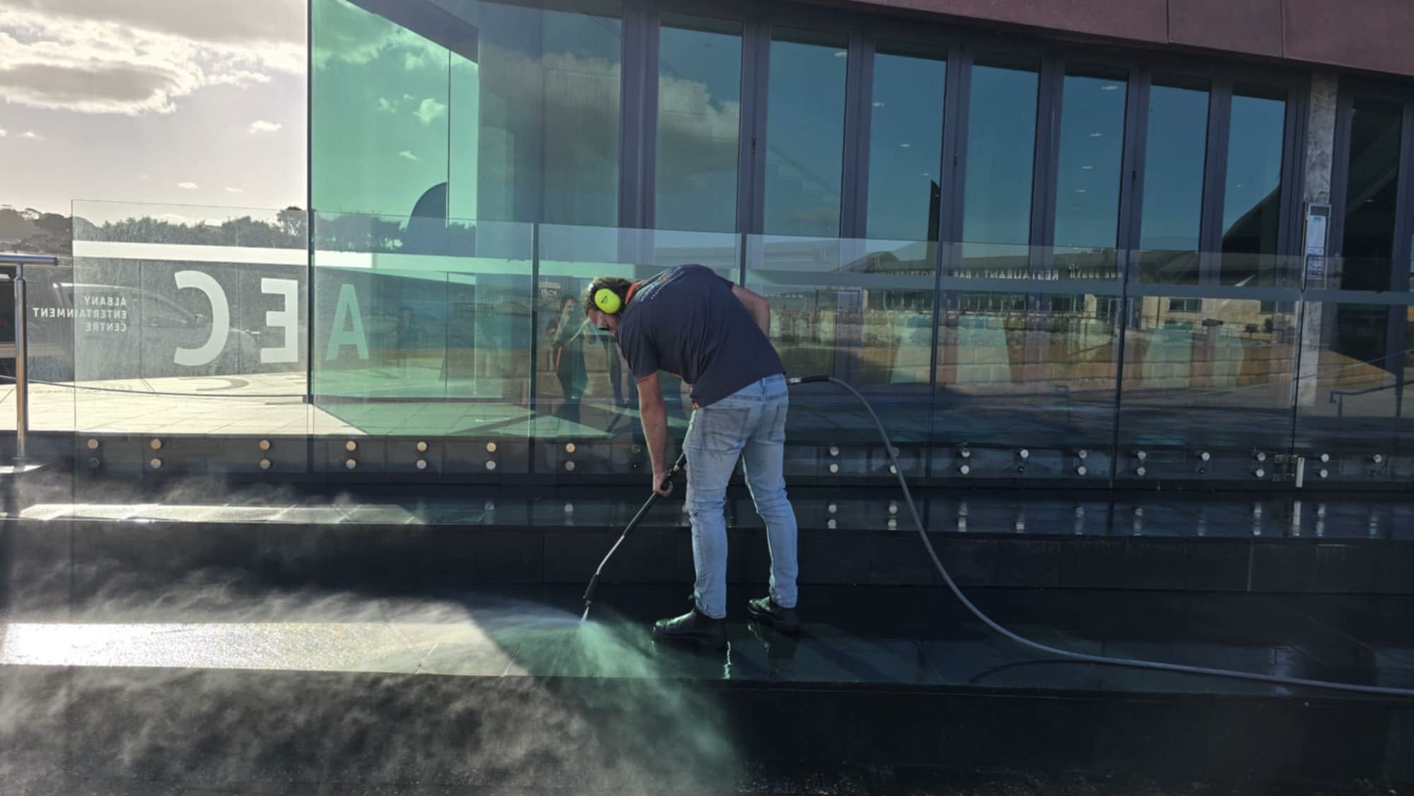 Worker cleaning glass exterior of modern building with high-pressure water hose, reflection of person in glass, blue sky with clouds in background.