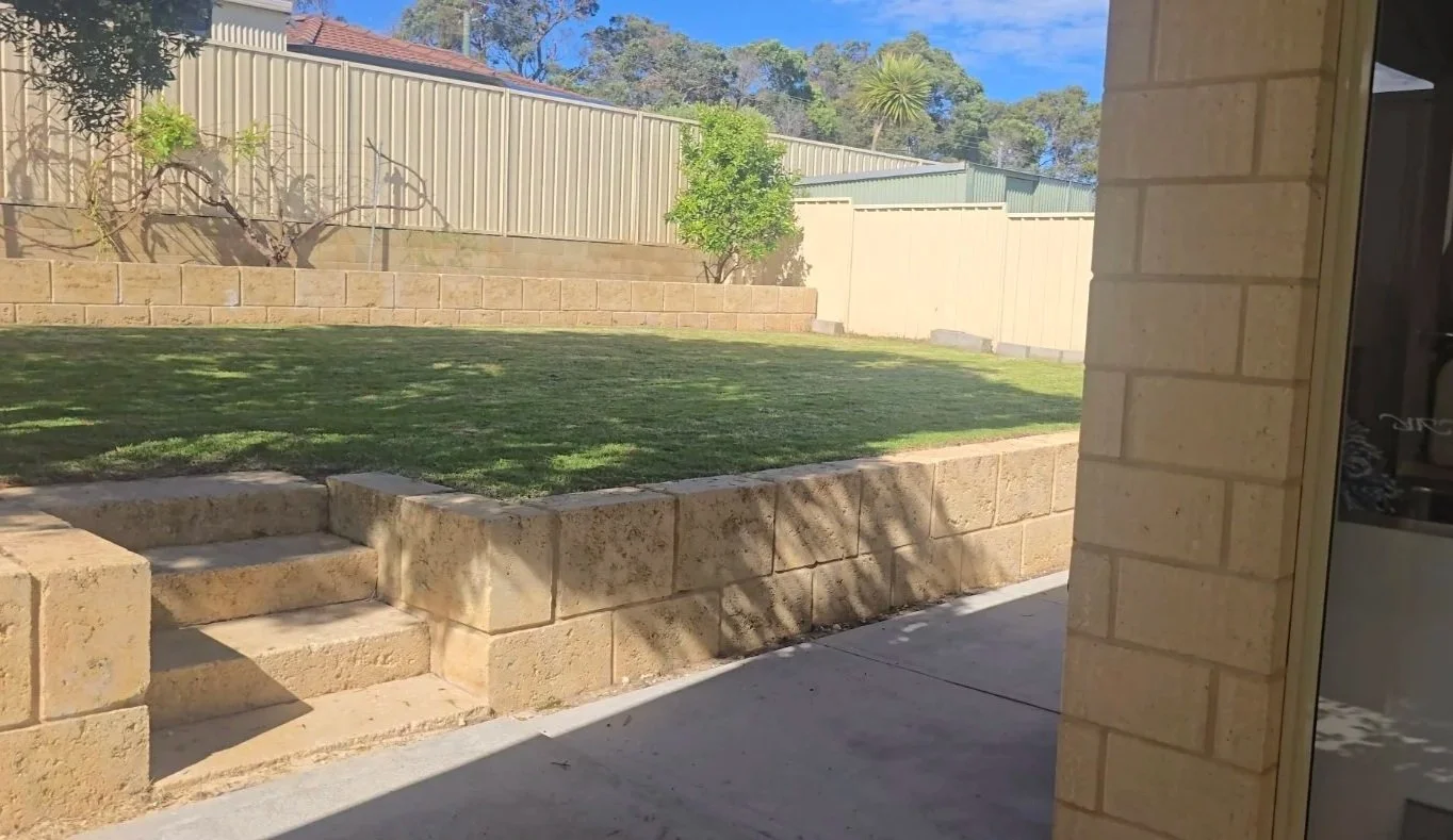 View of backyard with green grass, small trees, and a beige brick retaining wall under a clear blue sky, viewed from covered patio area.